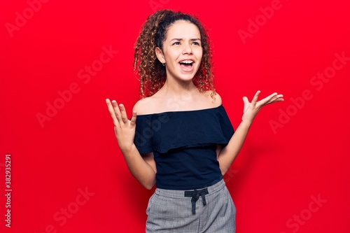 Beautiful kid girl with curly hair wearing casual clothes crazy and mad shouting and yelling with aggressive expression and arms raised. frustration concept.