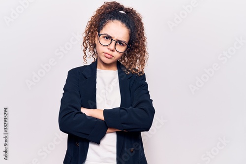 Beautiful kid girl with curly hair wearing business clothes and glasses skeptic and nervous, disapproving expression on face with crossed arms. negative person.