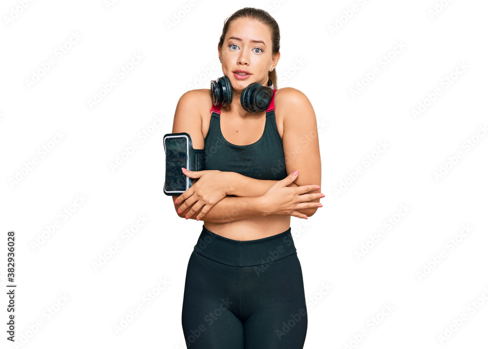 Beautiful blonde woman wearing gym clothes and using headphones shaking and freezing for winter cold with sad and shock expression on face Beautiful blonde woman wearing gym clothes and using headphones shaking and freezing for winter cold with sad and shock expression on face
