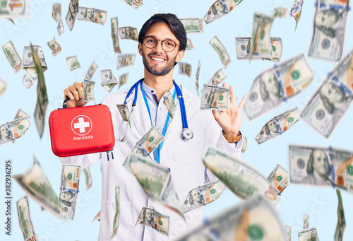 Handsome hispanic man wearing doctor coat holding first aid kit doing ok sign with fingers, smiling friendly gesturing excellent symbol