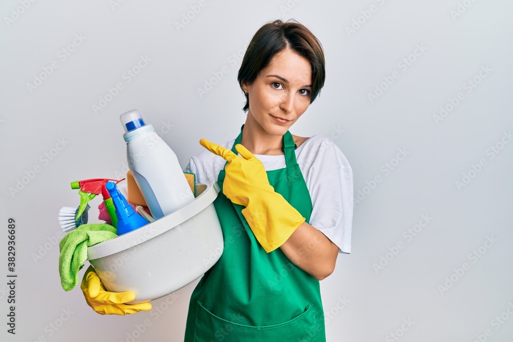 Young brunette woman with short hair wearing apron holding cleaning products pointing with hand finger to the side showing advertisement, serious and calm face Young brunette woman with short hair wearing apron holding cleaning products pointing with hand finger to the side showing advertisement, serious and calm face