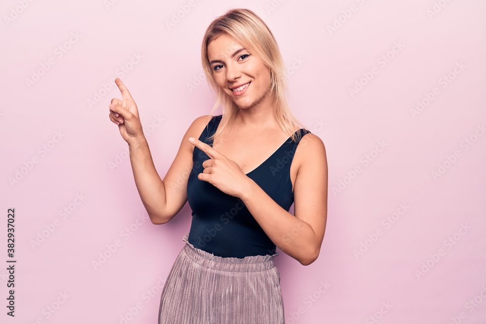 Young beautiful blonde woman wearing casual black t-shirt standing over pink background smiling and looking at the camera pointing with two hands and fingers to the side. Young beautiful blonde woman wearing casual black t-shirt standing over pink background smiling and looking at the camera pointing with two hands and fingers to the side.