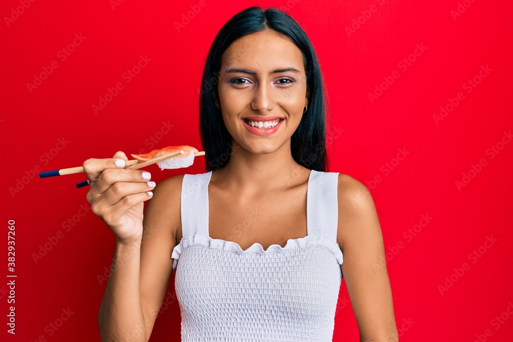 Young brunette woman eating prawn sushi using chopsticks looking positive and happy standing and smiling with a confident smile showing teeth Young brunette woman eating prawn sushi using chopsticks looking positive and happy standing and smiling with a confident smile showing teeth