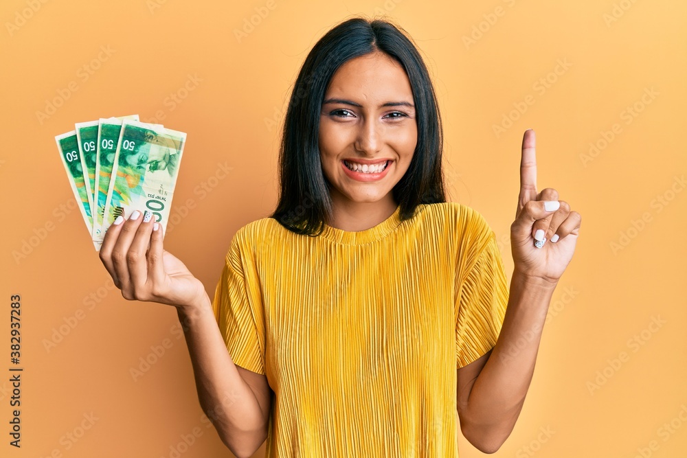Young brunette woman holding 50 israel shekels banknotes smiling with an idea or question pointing finger with happy face, number one Young brunette woman holding 50 israel shekels banknotes smiling with an idea or question pointing finger with happy face, number one