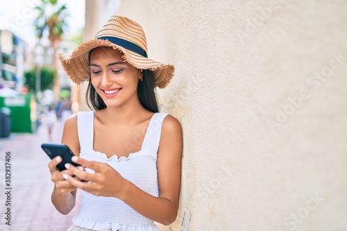 Young latin girl wearing summer style using smartphone leaning on the wall.