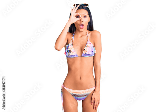 Young beautiful latin girl wearing bikini and sunglasses doing ok gesture shocked with surprised face, eye looking through fingers. unbelieving expression.