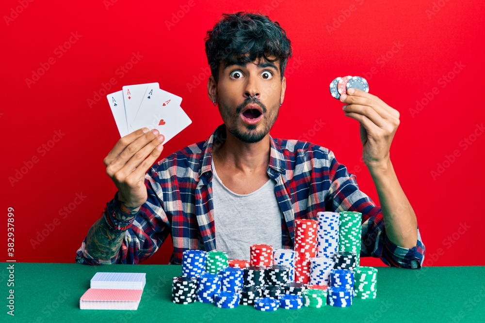 Young hispanic man playing poker holding cards and casino chips afraid and shocked with surprise and amazed expression, fear and excited face. Young hispanic man playing poker holding cards and casino chips afraid and shocked with surprise and amazed expression, fear and excited face.