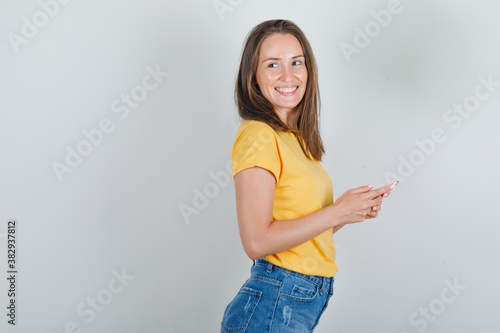 Young woman in t-shirt, shorts looking away with smartphone in hands and looking cheery .