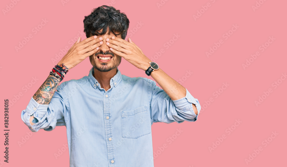 Young hispanic man wearing casual clothes covering eyes with hands smiling cheerful and funny. blind concept. Young hispanic man wearing casual clothes covering eyes with hands smiling cheerful and funny. blind concept.
