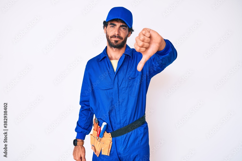 Handsome young man with curly hair and bear weaing handyman uniform looking unhappy and angry showing rejection and negative with thumbs down gesture. bad expression.