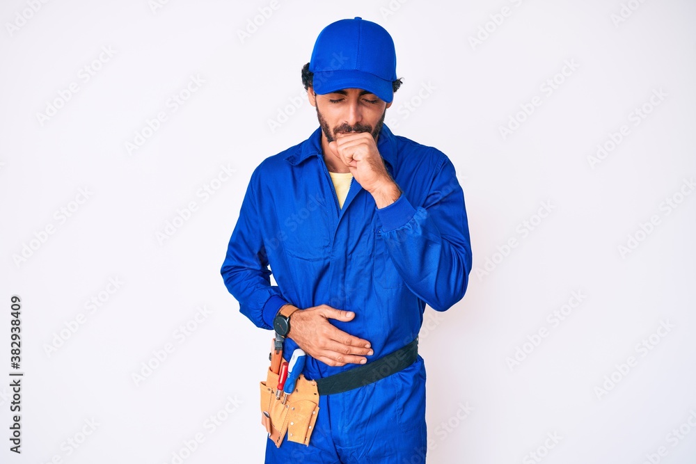 Handsome young man with curly hair and bear weaing handyman uniform feeling unwell and coughing as symptom for cold or bronchitis. health care concept.