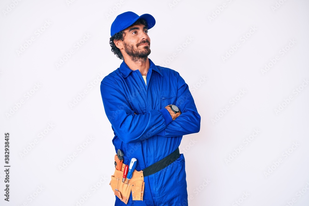 Handsome young man with curly hair and bear weaing handyman uniform looking to the side with arms crossed convinced and confident Handsome young man with curly hair and bear weaing handyman uniform looking to the side with arms crossed convinced and confident