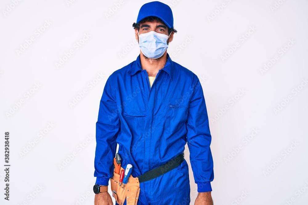 Handsome young man with curly hair and bear wearing handyman uniform and covid-19 safety mask with a happy and cool smile on face. lucky person. Handsome young man with curly hair and bear wearing handyman uniform and covid-19 safety mask with a happy and cool smile on face. lucky person.