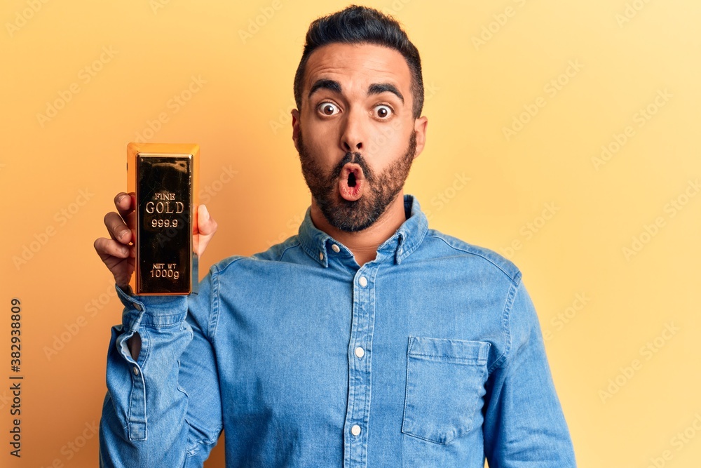 Young hispanic man holding gold ingot scared and amazed with open mouth ...