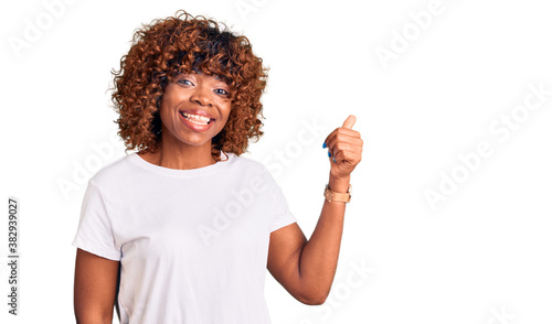 Young african american woman wearing casual white tshirt smiling with happy face looking and pointing to the side with thumb up.