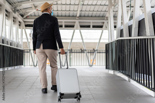 Casual businessman travel with trolley bag walking in the airport terminal. Young man wearing mask arrive to airport. Safety travel concept..