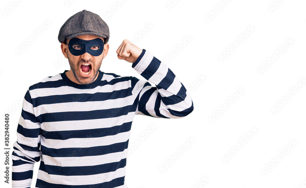 Young handsome man wearing burglar mask angry and mad raising fist frustrated and furious while shouting with anger. rage and aggressive concept. Young handsome man wearing burglar mask angry and mad raising fist frustrated and furious while shouting with anger. rage and aggressive concept.