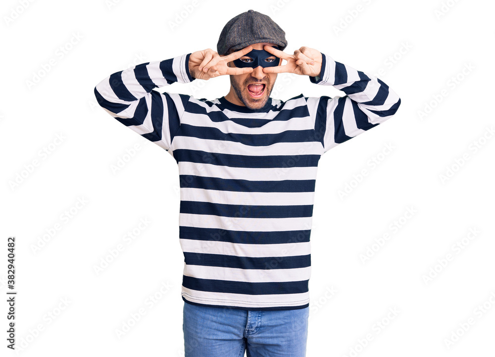 Young handsome man wearing burglar mask doing peace symbol with fingers over face, smiling cheerful showing victory Young handsome man wearing burglar mask doing peace symbol with fingers over face, smiling cheerful showing victory