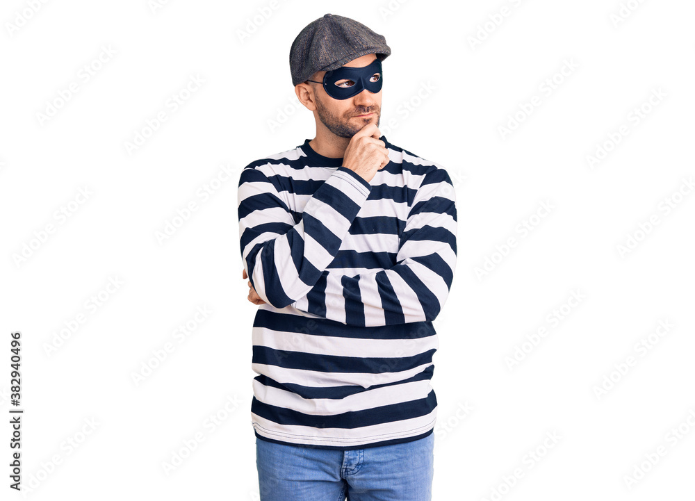 Young handsome man wearing burglar mask with hand on chin thinking about question, pensive expression. smiling with thoughtful face. doubt concept.