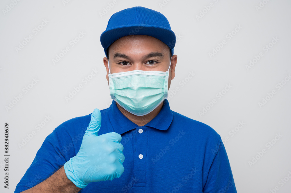 Blue delivery asian man wearing protection mask and medical rubber gloves standing showing thumbs up on isolated white background safety deliver concept. Blue delivery asian man wearing protection mask and medical rubber gloves standing showing thumbs up on isolated white background safety deliver concept.
