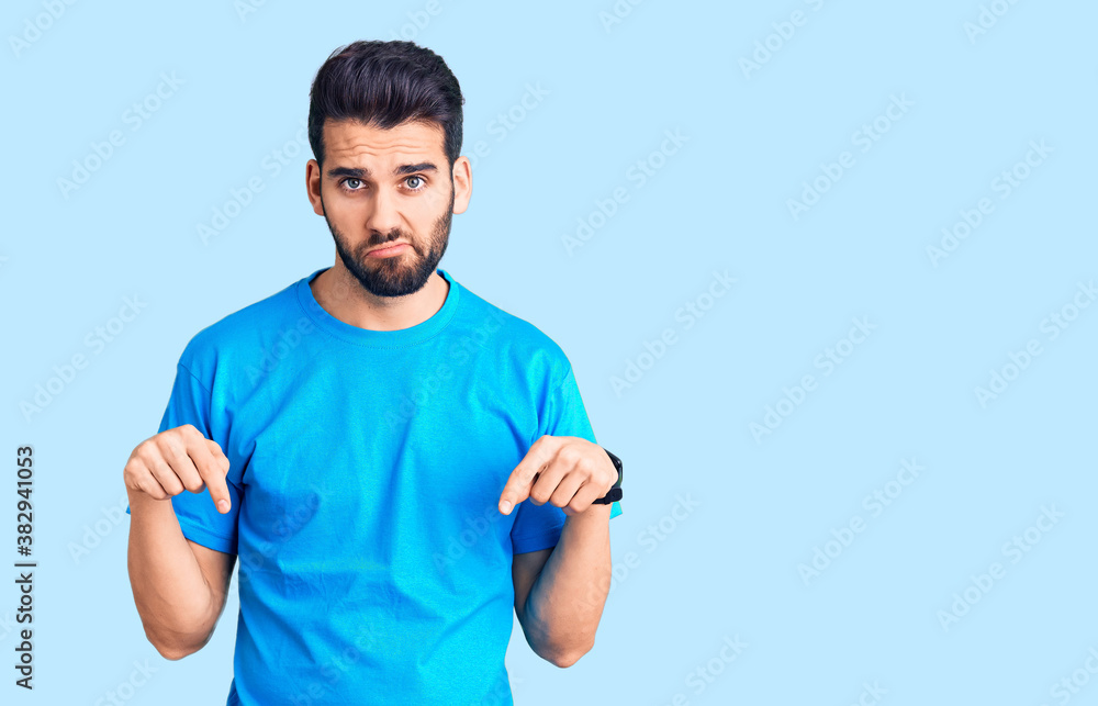 Young handsome man with beard wearing casual t-shirt pointing down looking sad and upset, indicating direction with fingers, unhappy and depressed. Young handsome man with beard wearing casual t-shirt pointing down looking sad and upset, indicating direction with fingers, unhappy and depressed.