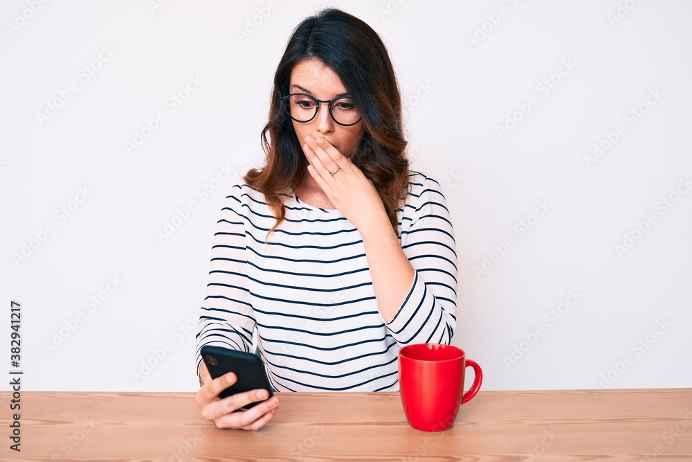 Young beautiful brunette woman using smartphone covering mouth with hand, shocked and afraid for mistake. surprised expression Young beautiful brunette woman using smartphone covering mouth with hand, shocked and afraid for mistake. surprised expression