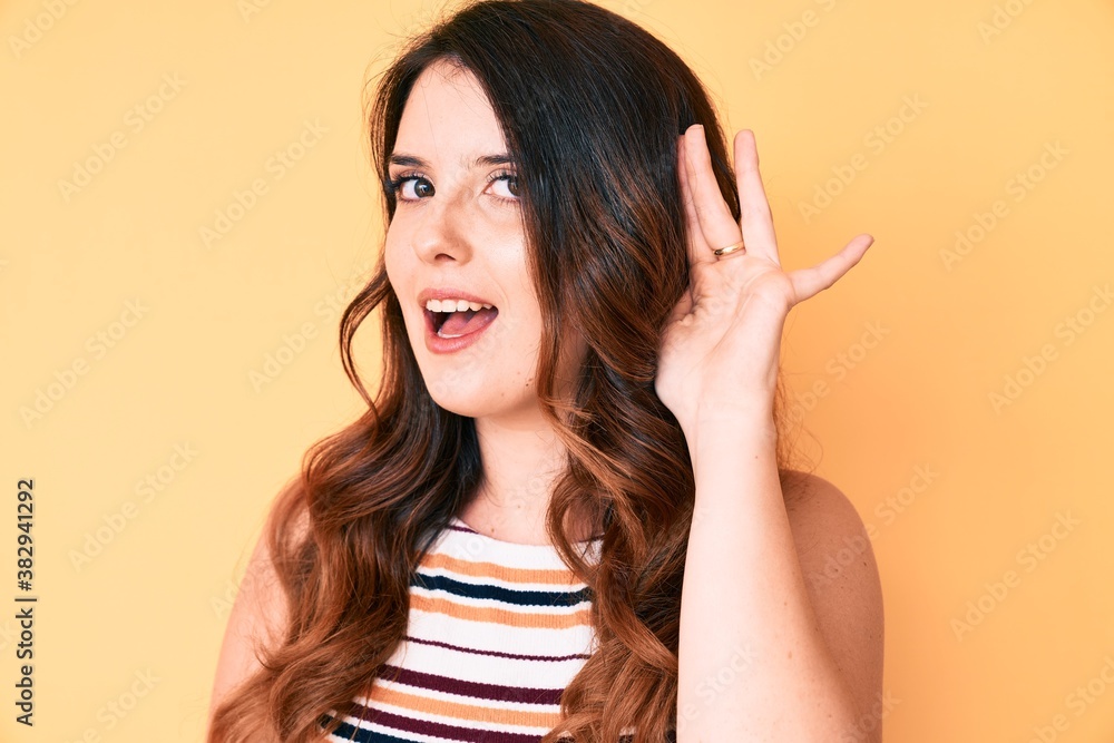 Young beautiful brunette woman over yellow background smiling with hand over ear listening and hearing to rumor or gossip. deafness concept. Young beautiful brunette woman over yellow background smiling with hand over ear listening and hearing to rumor or gossip. deafness concept.