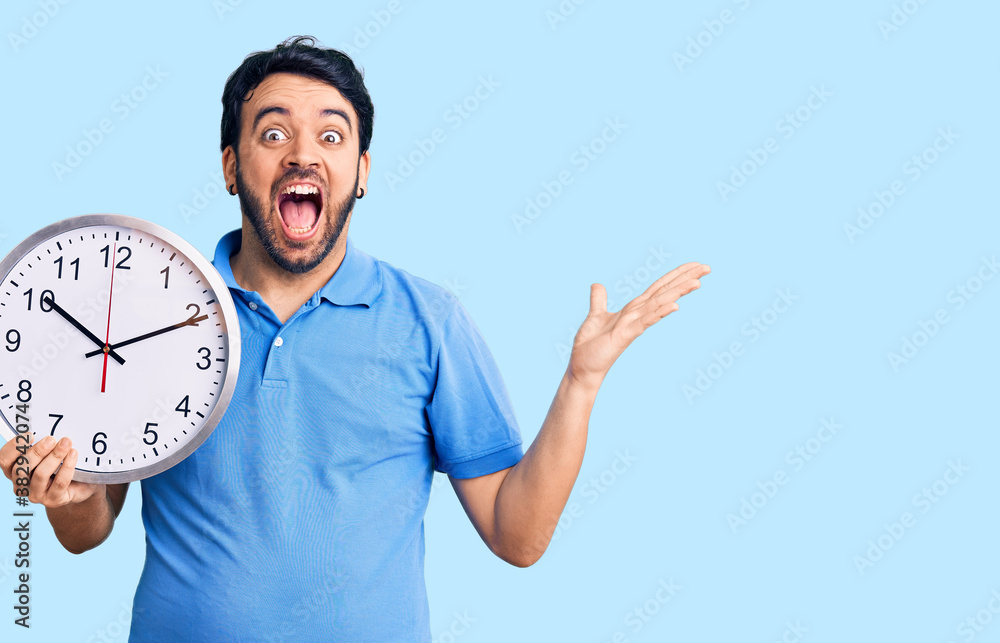 Young hispanic man holding big clock celebrating victory with happy smile and winner expression with raised hands Young hispanic man holding big clock celebrating victory with happy smile and winner expression with raised hands