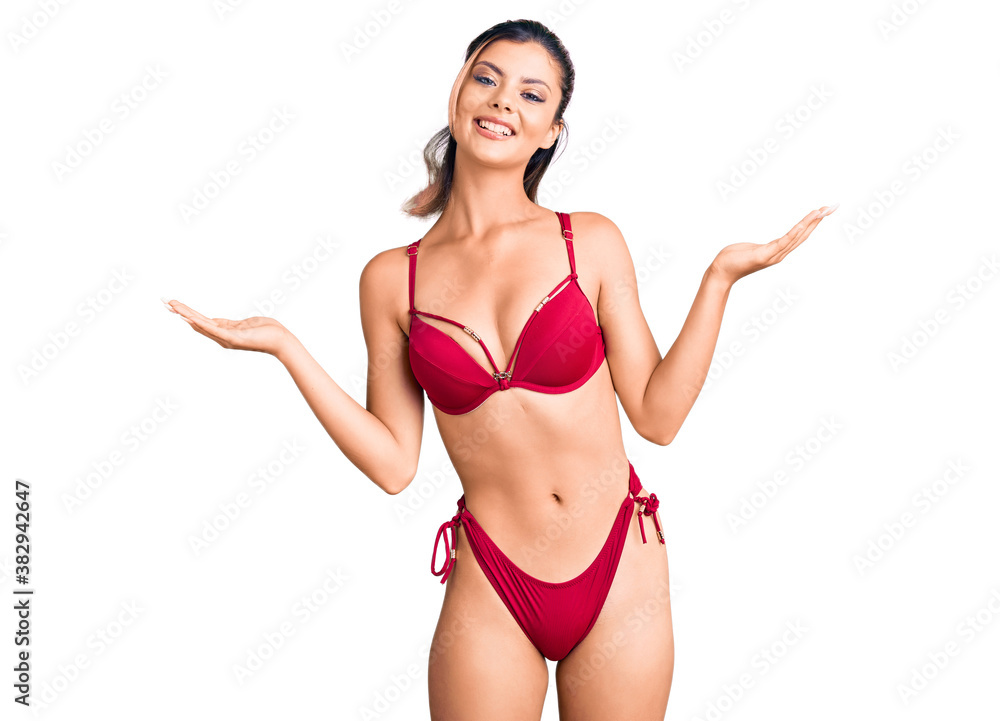 Young beautiful woman wearing bikini smiling showing both hands open palms, presenting and advertising comparison and balance Young beautiful woman wearing bikini smiling showing both hands open palms, presenting and advertising comparison and balance