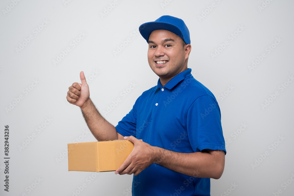 Young smiling asian delivery man in blue uniform holding box parcel cardboard and showing thumbs up on isolated white background. Young smiling asian delivery man in blue uniform holding box parcel cardboard and showing thumbs up on isolated white background.