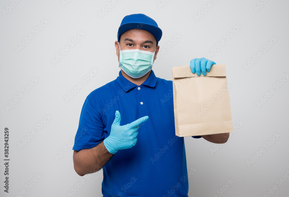 Young asian delivery man in blue uniform wearing protection mask and medical rubber gloves giving the paper bag to customer on isolated white background. Safety deliver concept. Young asian delivery man in blue uniform wearing protection mask and medical rubber gloves giving the paper bag to customer on isolated white background. Safety deliver concept.