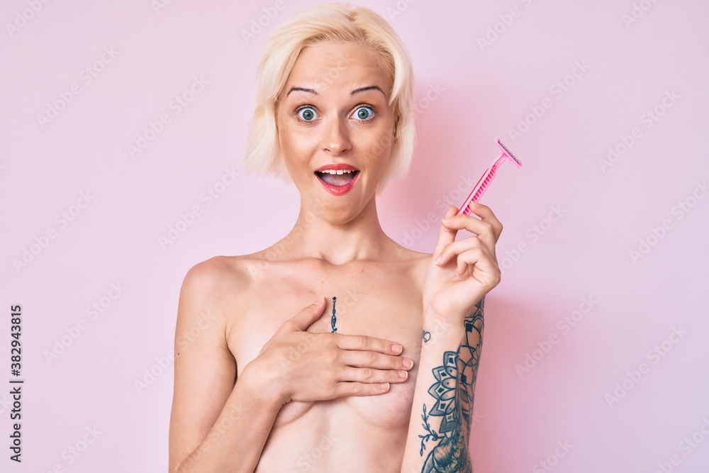 Obraz premium Young blonde woman with tattoo standing shirtless holding razor celebrating crazy and amazed for success with open eyes screaming excited.
