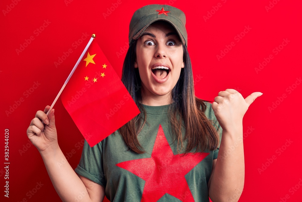 Beautiful patriotic woman wearing t-shirt with red star communist symbol holding china flag pointing thumb up to the side smiling happy with open mouth Beautiful patriotic woman wearing t-shirt with red star communist symbol holding china flag pointing thumb up to the side smiling happy with open mouth