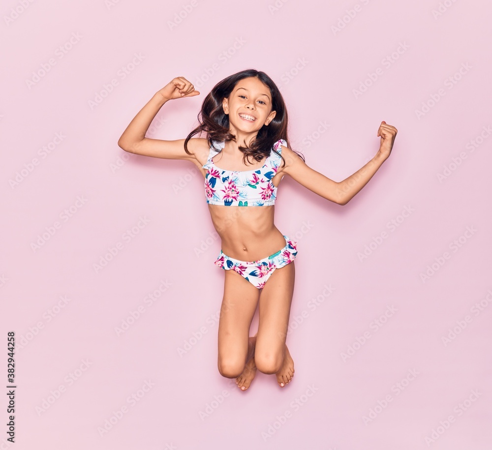 Naklejka premium Adorable hispanic child girl on vacation wearing bikini smiling happy. Jumping with smile on face over isolated pink background