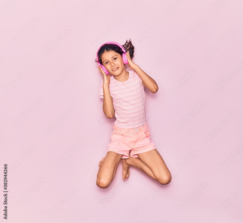 Adorable hispanic child girl smiling happy. Jumping with smile on face listening to music using pink headphones over isolated background Adorable hispanic child girl smiling happy. Jumping with smile on face listening to music using pink headphones over isolated background