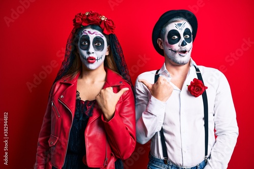 Couple wearing day of the dead costume over red surprised pointing with hand finger to the side, open mouth amazed expression.