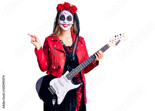 Woman wearing day of the dead costume playing electric guitar smiling happy pointing with hand and finger to the side