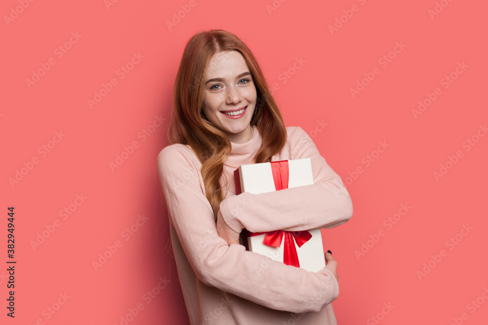 Charming red haired woman with freckles smiling at camera while embracing a present in a warm sweater posing on a red wall Charming red haired woman with freckles smiling at camera while embracing a present in a warm sweater posing on a red wall
