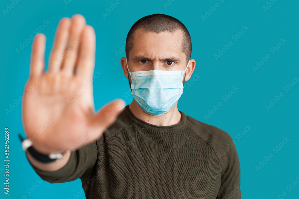 Close up photo of a caucasian man in sweater wearing a medical mask on a blue studio wall gesturing stop and rejection sign with palm Close up photo of a caucasian man in sweater wearing a medical mask on a blue studio wall gesturing stop and rejection sign with palm