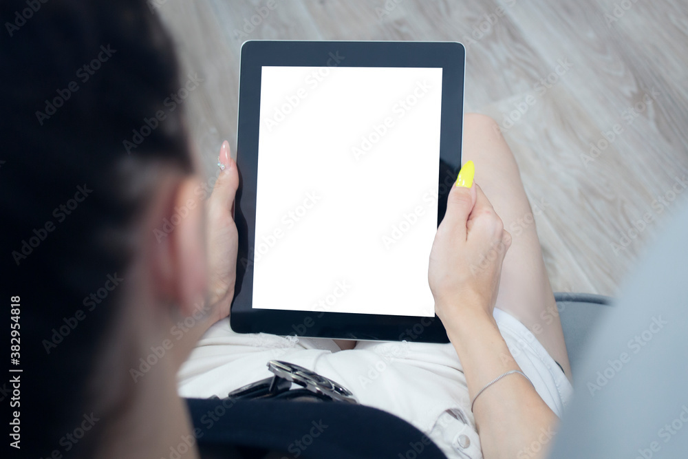 The girl sits in a chair and holds a tablet with a white screen in her hands. Mockup technology.