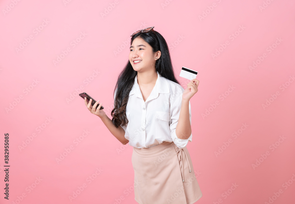 Young smiling Asian woman holding credit card with smartphone and looking to copy space on pink background. Young smiling Asian woman holding credit card with smartphone and looking to copy space on pink background.