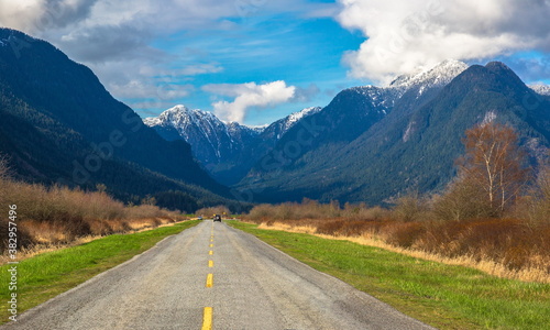 A country road in the Pitt River Valley runs through farm fields and forest to Pitt Lake on the background of the snow-covered mountain range and a beautiful cloudy sky