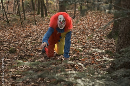 Halloween holiday. Horror and fear. Scary aggressive clown with red hair in the autumn dark forest. Masquerade and carnival. Scary clown costumes. Creepy clown in the autumn forest
