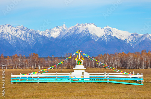Buddhist stupa decorated with prayer flags against the background of snow-capped peaks of the Eastern Sayan Mountains on a sunny autumn day. Siberia. Baikal region. Buryatia. Tunka valley