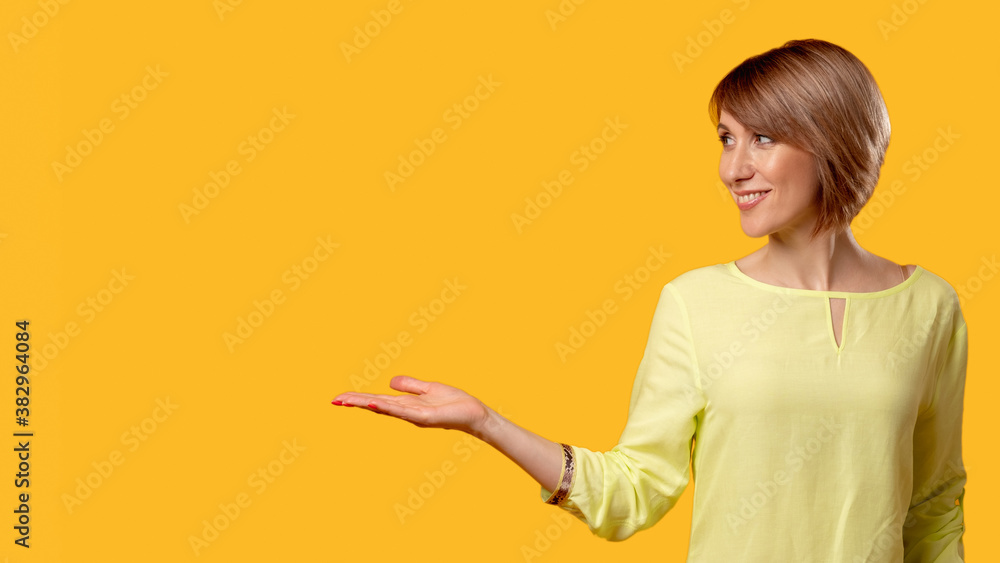 Special offer. Solution way. Advertising background. Cheerful mature woman in yellow blouse recommending virtual invisible product with open palm smiling isolated on orange empty space. Special offer. Solution way. Advertising background. Cheerful mature woman in yellow blouse recommending virtual invisible product with open palm smiling isolated on orange empty space.