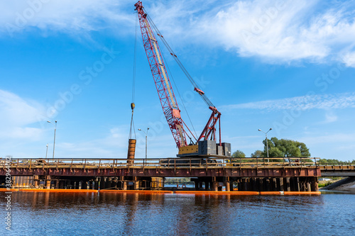 Construction of a new bridge. A crane on the background of a blue sky above the lake lifts a metal pipe