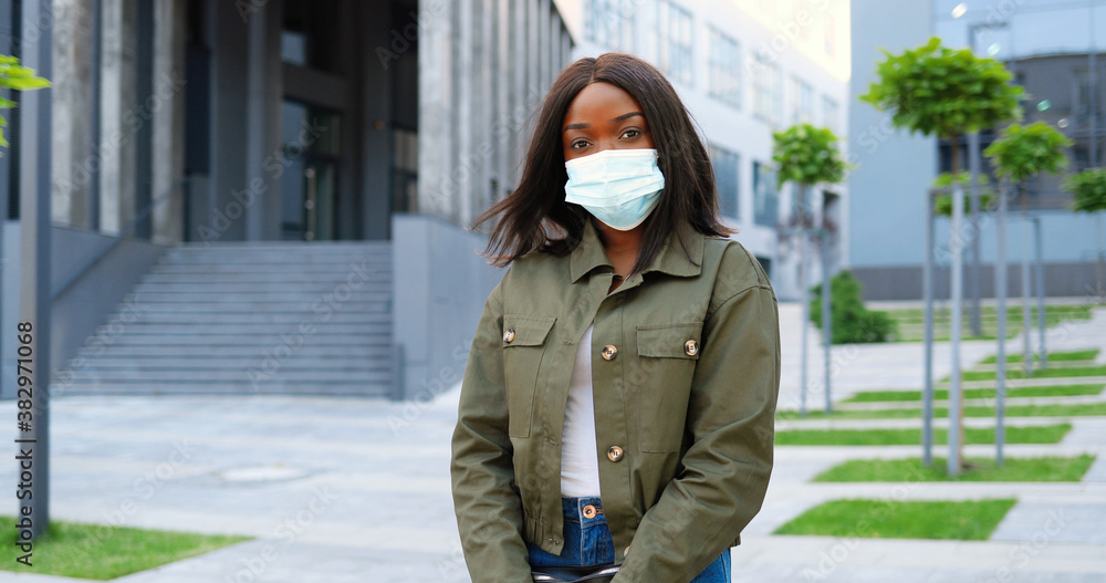 Portrait of African American young stylish pretty woman in medical mask looking at camera and standing at urban street. Beautiful female outdoors in city during pandemic. Coronavirus lockdown.