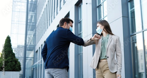 Caucasian young joyful female and male in medical masks meeting at street and greeting with elbows. Happy businessman and businesswoman talking and chatting cheerfully. Couple of friends.
