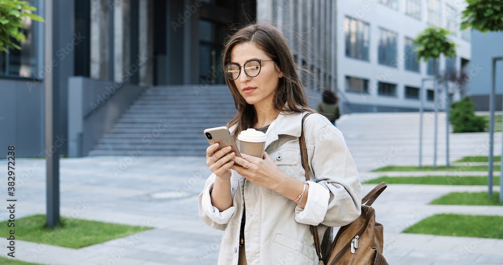 Caucasian cheerful young stylish woman tapping or scrolling on smartphone and sipping hot drink in the morning at street. Beautiful happy female texting message on phone and drinking coffee. Outside.