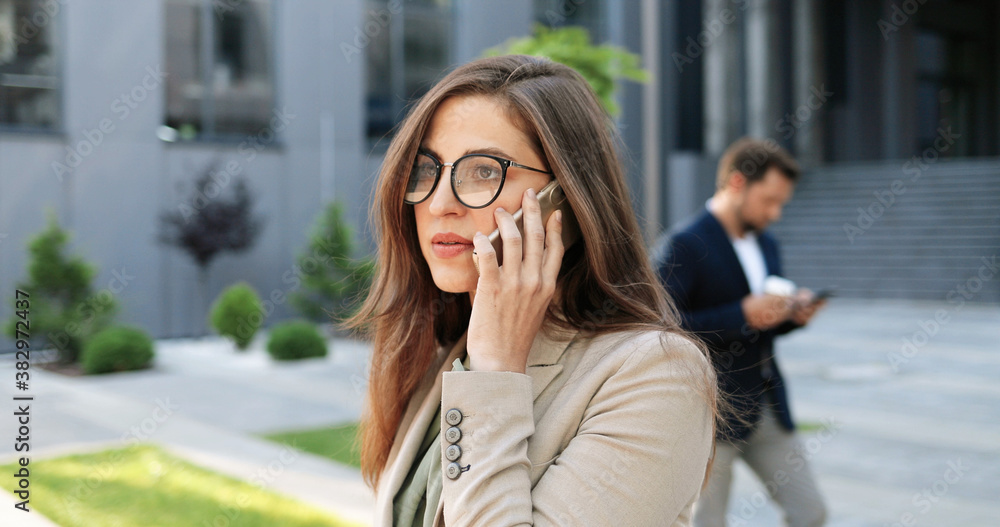 Caucasian young stylish businesswoman in glasses talking on cellphone at street. Beautiful female speaking on mobile phone outdoors. Attractive woman in eyeglasses having telephone call. Conversation.